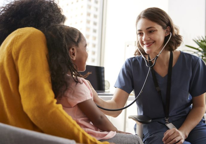 Female Pediatrician Wearing Scrubs Listening To Girls Chest With Stethoscope In Hospital Office